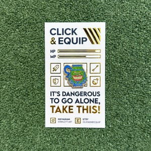 May include: A green and blue enamel pin featuring a grinning face inside a pitcher. The pin has the text "CLICK & EQUIP" and "IT'S DANGEROUS TO GO ALONE, TAKE THIS!" The pin also has the text "INSTAGRAM STANLEYT.ART" and "ETSY CLICKANDEQUIP".