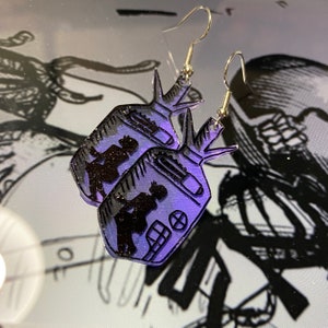 May include: A pair of purple and black earrings with a silhouette of two people in a vintage television set. The earrings are made of acrylic and have silver hooks.