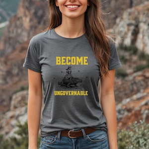 Puede incluir: Una camiseta gris con las palabras "BECOME UNGOVERNABLE" en amarillo. La camiseta presenta un gr&aacute;fico de una persona montando un caim&aacute;n. La camiseta tiene mangas cortas y cuello redondo.