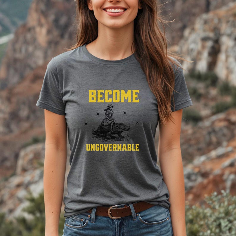 Puede incluir: Una camiseta gris con las palabras "BECOME UNGOVERNABLE" en amarillo. La camiseta presenta un gr&aacute;fico de una persona montando un caim&aacute;n. La camiseta tiene mangas cortas y cuello redondo.
