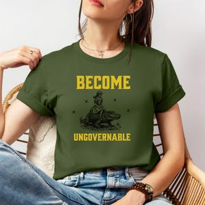 Puede incluir: Camiseta verde oliva con las palabras "BECOME UNGOVERNABLE" en letras amarillas. Debajo, una ilustraci&oacute;n negra de una persona montando un caim&aacute;n. La camiseta es de manga corta.