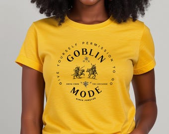 May include: Mustard yellow t-shirt with rolled sleeves. The shirt features a circular graphic with the words "GOBLIN MODE" and the text "GIVE YOURSELF PERMISSION TO GO" and "DRINK FROM THE CONTAINER" in a black font. The shirt also has a graphic of two figures on horseback.