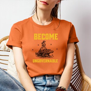 Puede incluir: Camiseta naranja con mangas enrolladas. El texto amarillo dice "BECOME UNGOVERNABLE". Un gr&aacute;fico muestra a una persona montando un caim&aacute;n. La camiseta est&aacute; hecha de un material suave.