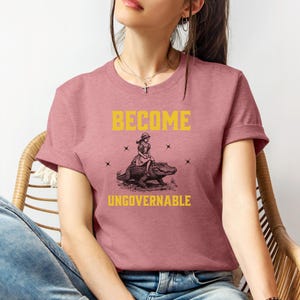 Puede incluir: Camiseta rosa polvoriento con las palabras "BECOME UNGOVERNABLE" en amarillo. La camiseta presenta un gr&aacute;fico de una persona montando un caim&aacute;n. El dise&ntilde;o est&aacute; centrado en la parte delantera de la camiseta.