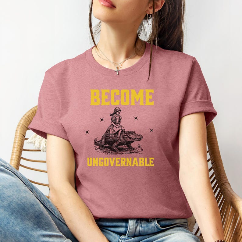 Puede incluir: Camiseta rosa polvoriento con las palabras "BECOME UNGOVERNABLE" en amarillo. La camiseta presenta un gr&aacute;fico de una persona montando un caim&aacute;n. El dise&ntilde;o est&aacute; centrado en la parte delantera de la camiseta.