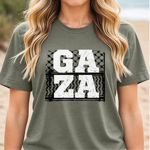 May include: Olive green t-shirt with the word "GAZA" in large white block letters. The letters are set against a black and white patterned background, creating a bold graphic design.