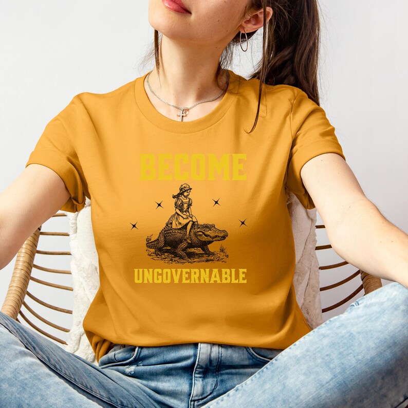 Puede incluir: Camiseta de color amarillo mostaza con las palabras "BECOME UNGOVERNABLE" en letras amarillas. La camiseta presenta un gr&aacute;fico de una persona montando un caim&aacute;n. La persona lleva un vestido y un sombrero. El caim&aacute;n es marr&oacute;n.