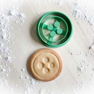 Könnte beinhalten: Ein grüner Plastik-Ausstecher in Form eines Knopfes mit vier kleineren Kreisen darin. Neben dem Ausstecher liegt ein Keks mit Knopfdesign.