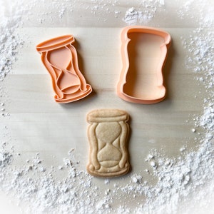 Peut inclure: Deux emportes-pièces en plastique orange en forme de sablier, l'un avec un design détaillé et l'autre avec un contour simple, et un biscuit cuit en forme de sablier.