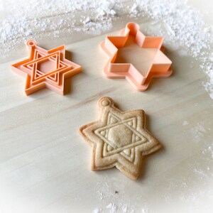 Op de afbeelding: Drie koekjesvormen in de vorm van een zespuntige ster, twee zijn oranje en &eacute;&eacute;n is beige. Een koekje in de vorm van een zespuntige ster ligt op een houten oppervlak.