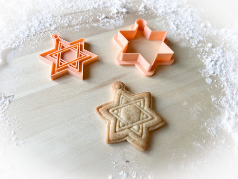 Op de afbeelding: Drie koekjesvormen in de vorm van een zespuntige ster, twee zijn oranje en &eacute;&eacute;n is beige. Een koekje in de vorm van een zespuntige ster ligt op een houten oppervlak.