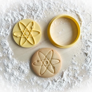 May include: A yellow plastic cookie cutter shaped like an atom, with a cookie cut out using the cutter on a white surface.