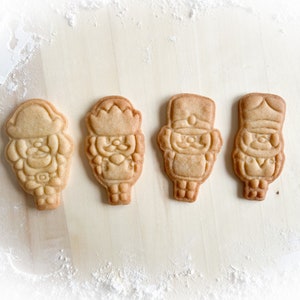 Könnte beinhalten: Vier Lebkuchen in Form von Nussknackern, verziert mit Zuckerguss und Details. Die Kekse sind in einer Reihe auf einer hellbraunen Oberfläche angeordnet.