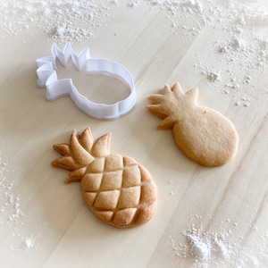 Peut inclure: Deux biscuits en forme d'ananas et un emporte-pièce en plastique blanc en forme d'ananas sur une surface en bois clair avec de la poudre blanche saupoudrée dessus.