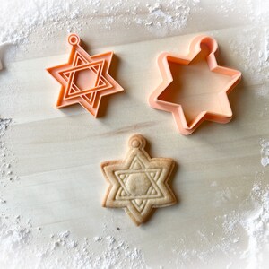 Op de afbeelding: Twee oranje plastic koekjesvormen in de vorm van een zespuntige ster, en een gebakken koekje in de vorm van een zespuntige ster.