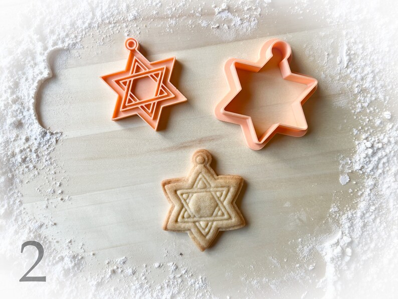 Op de afbeelding: Twee oranje plastic koekjesvormen in de vorm van een zespuntige ster, en een gebakken koekje in de vorm van een zespuntige ster.