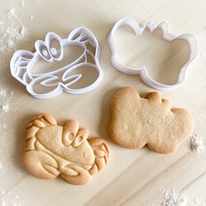 May include: Two white plastic cookie cutters shaped like crabs, one with a simple design and the other with a more detailed design, and a baked cookie shaped like a crab.