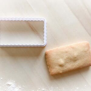 Peut inclure: Un emporte-pièce en plastique blanc en forme de rectangle avec des bords festonnés. Un biscuit nature brun clair se trouve à côté de l'emporte-pièce.