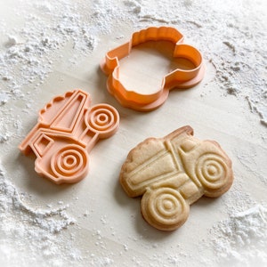 May include: A set of orange plastic cookie cutters and a baked cookie, all in the shape of a dump truck, are arranged on a surface dusted with flour. The cookie cutter is in two pieces. The baked cookie has detailed lines, showing the truck's features.