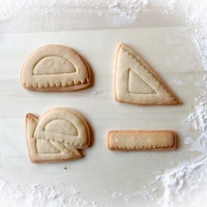 May include: Four homemade sugar cookies shaped like protractors and rulers. The cookies are on a light brown surface covered in white powdered sugar.