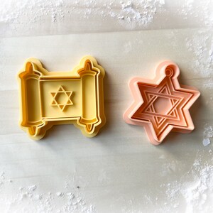 Op de afbeelding: Twee gele plastic koekjesvormen, een in de vorm van een Davidster en de andere in de vorm van een Thora-rol met een Davidster erin.