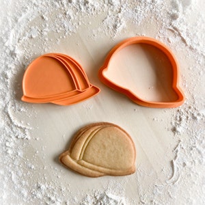 Könnte beinhalten: Zwei orangefarbene Plastik-Ausstecher in Form von Bauhelmen, einer mit einem Keks darin. Der Keks ist hellbraun und hat eine ähnliche Form wie die Ausstecher.