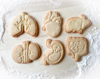 Puede incluir: Un conjunto de siete galletas de azúcar con forma de órganos humanos, que incluyen un corazón, pulmones, riñones, estómago, intestinos e hígado. Las galletas son de color marrón claro y tienen diseños detallados.