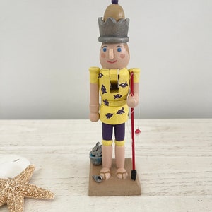 Pode incluir: Uma figura de quebra-nozes de madeira vestida com uma camisa amarela com peixes roxos, cal&ccedil;&otilde;es roxos e uma coroa cinzenta. O quebra-nozes segura uma vara de pesca vermelha com um boia vermelha.