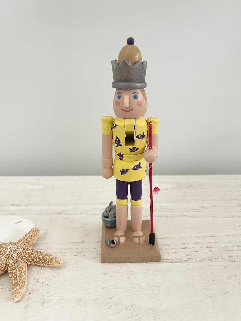 Pode incluir: Uma figura de quebra-nozes de madeira vestida com uma camisa amarela com peixes roxos, cal&ccedil;&otilde;es roxos e uma coroa cinzenta. O quebra-nozes segura uma vara de pesca vermelha com um boia vermelha.