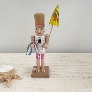 Pode incluir: Uma figura de quebra-nozes de madeira vestida com um mai&ocirc; de bolinhas rosa e branco, segurando um guarda-chuva de praia amarelo e vermelho. O quebra-nozes est&aacute; em p&eacute; sobre uma base marrom.