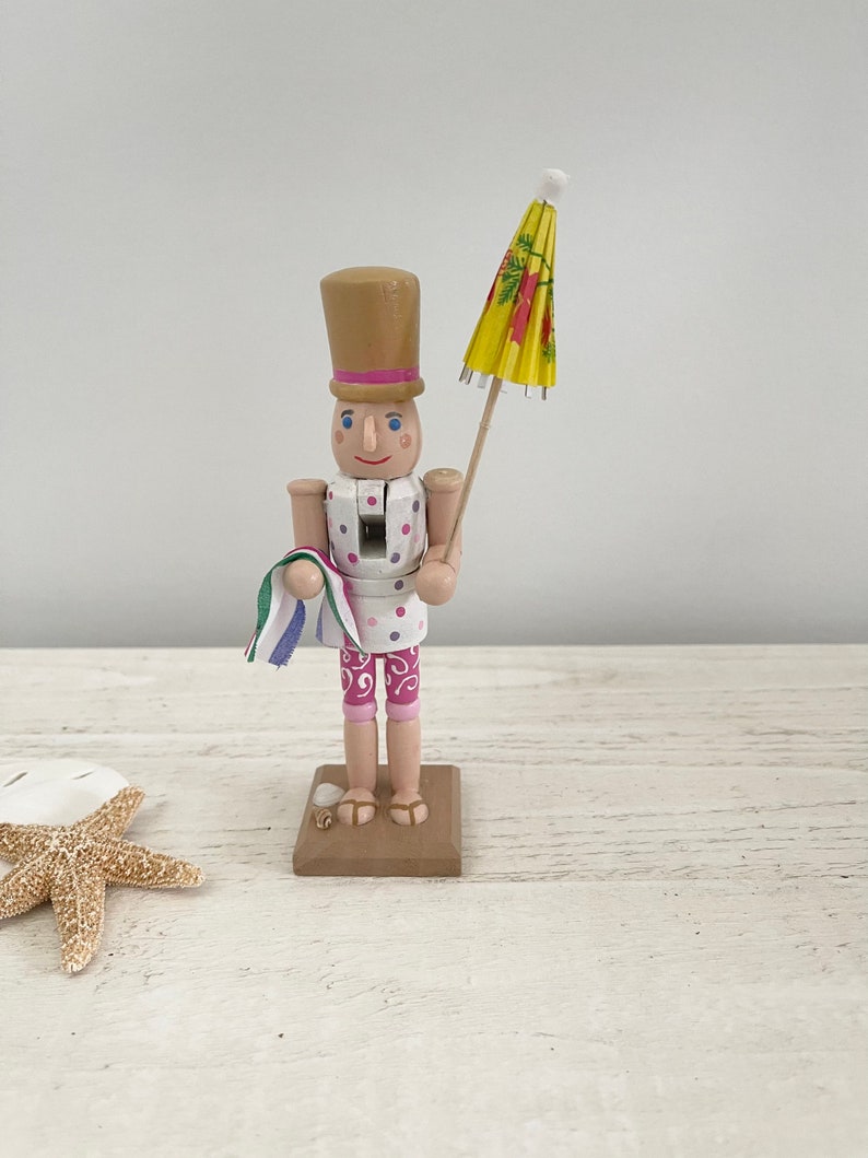 Pode incluir: Uma figura de quebra-nozes de madeira vestida com um mai&ocirc; de bolinhas rosa e branco, segurando um guarda-chuva de praia amarelo e vermelho. O quebra-nozes est&aacute; em p&eacute; sobre uma base marrom.
