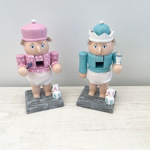 May include: Two wooden nutcracker figurines, one in pink and white and the other in blue and white, both dressed as babies with pacifiers and blocks.