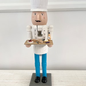 May include: A wooden chef nutcracker holding a tray with a baguette, cheese, grapes, and a bottle of wine. The nutcracker is wearing a white chef hat, a white chef coat, blue pants, and black shoes. The tray is brown wood.