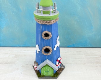May include: A blue and green wooden lighthouse with a white rope detail, two birdhouse openings, and a green door. The lighthouse is decorated with seashells, starfish, and a red and white life preserver.