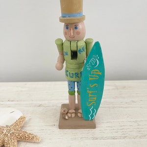 Pode incluir: Uma figura de quebra-nozes de madeira vestida de surfista. Ele est&aacute; usando uma camisa verde com a palavra "SURF" nela, sunga azul e um chap&eacute;u-coco marrom com uma faixa azul. Ele est&aacute; segurando uma prancha de surf azul com as palavras "Surf's Up" escritas nela.