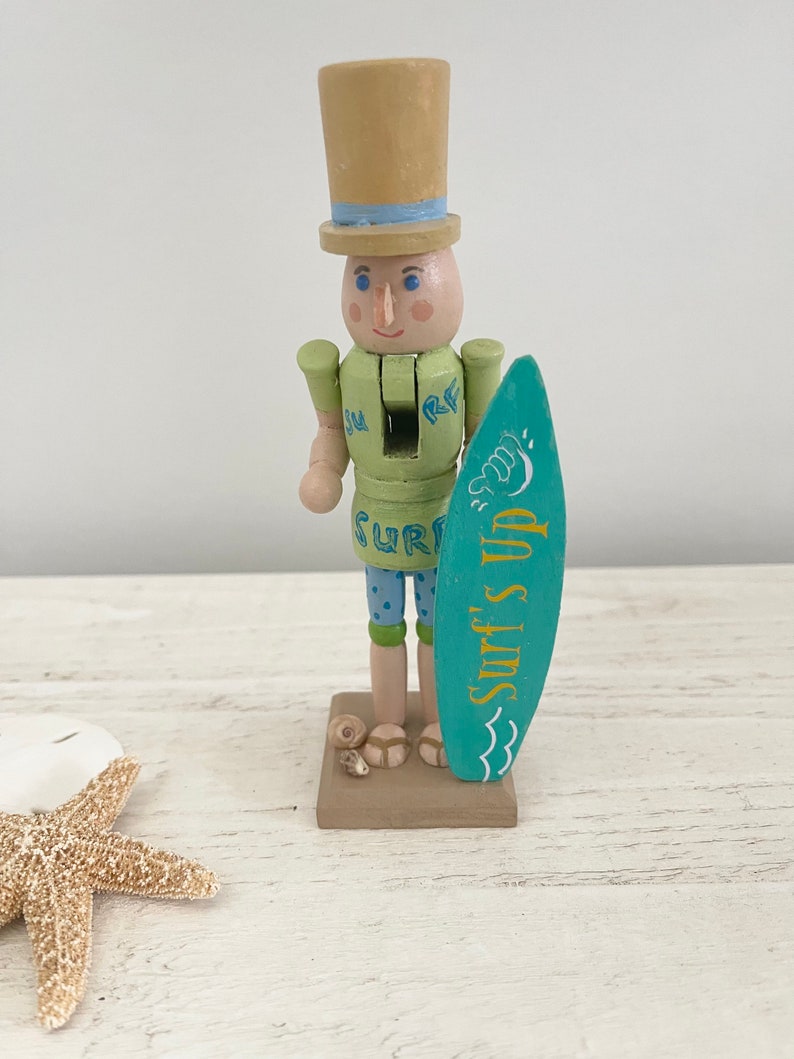 Pode incluir: Uma figura de quebra-nozes de madeira vestida de surfista. Ele est&aacute; usando uma camisa verde com a palavra "SURF" nela, sunga azul e um chap&eacute;u-coco marrom com uma faixa azul. Ele est&aacute; segurando uma prancha de surf azul com as palavras "Surf's Up" escritas nela.