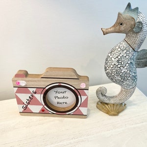 May include: A wooden camera-shaped photo frame with a pink and white geometric pattern. The frame has a small opening for a photo and the words "Your Photo Here" are printed inside. The frame is sitting on a white surface next to a decorative seahorse figurine.