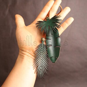 Pode incluir: Um conjunto de tr&ecirc;s pingentes de couro verde em forma de folhas de palmeira. Os pingentes s&atilde;o recortados em forma de folhas de palmeira e s&atilde;o presos a um cord&atilde;o de couro.