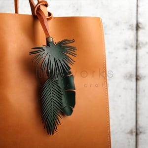 Pode incluir: Uma bolsa tote de couro marrom com um charme de folha de palmeira de couro verde preso &agrave; al&ccedil;a. O charme &eacute; feito de duas folhas, uma maior que a outra.