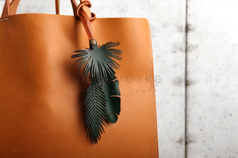 Pode incluir: Uma bolsa tote de couro marrom com um charme de folha de palmeira de couro verde preso &agrave; al&ccedil;a. O charme &eacute; feito de duas folhas, uma maior que a outra.