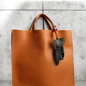 Pode incluir: Uma bolsa tote de couro marrom com um charme de folha de couro verde. A bolsa tem um design simples e &eacute; feita de couro de alta qualidade.
