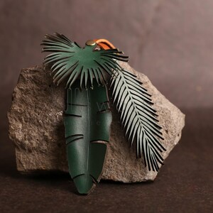 Pode incluir: Um amuleto de couro verde em forma de folha de palmeira e samambaia. As folhas s&atilde;o presas a um cord&atilde;o de couro marrom.