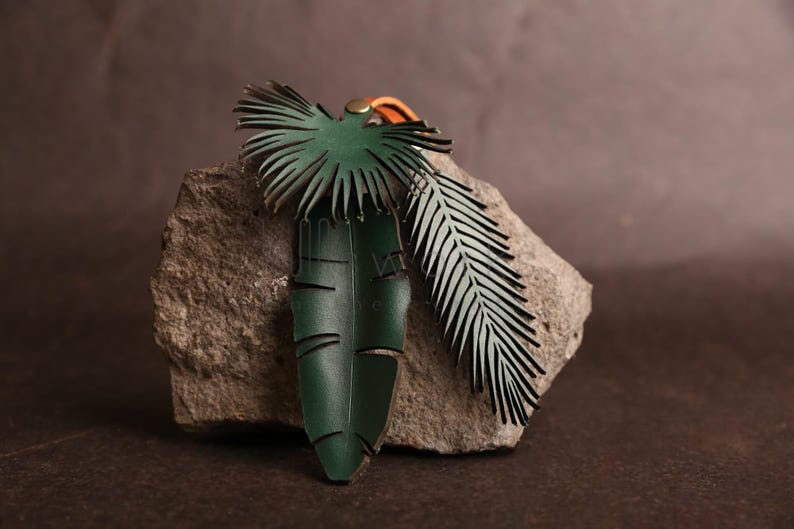 Pode incluir: Um amuleto de couro verde em forma de folha de palmeira e samambaia. As folhas s&atilde;o presas a um cord&atilde;o de couro marrom.