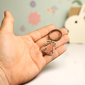 May include: A brown and white bunny keychain with a bronze-colored ring. The bunny is a simple silhouette design. The keychain is held in a hand.