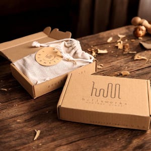 Pode incluir: Duas caixas de cart&atilde;o castanho com o texto "hool hillsworks" e "hillsworks" impresso nelas. A caixa &agrave; esquerda est&aacute; aberta e cont&eacute;m um saco de tecido branco com um r&oacute;tulo redondo que diz "hillsworks".