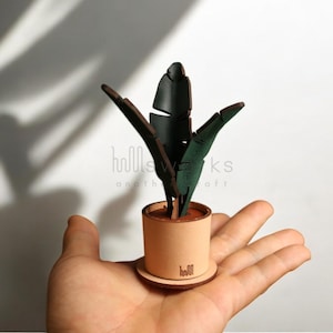May include: A small, handcrafted plant sculpture featuring dark green leaves and a light brown pot. The plant is in a round pot with a matching saucer. The sculpture is held in a hand, highlighting the craftsmanship and detail.