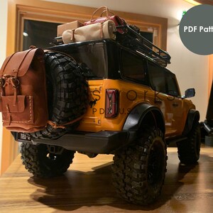 以下が含まれることがあります： 黒いルーフラックとスペアタイヤに茶色の革のバックパックが取り付けられた黄色のフォードブロンコモデルカー。車は木製の表面にあり、背面に「TRX-4」のステッカーが付いています。