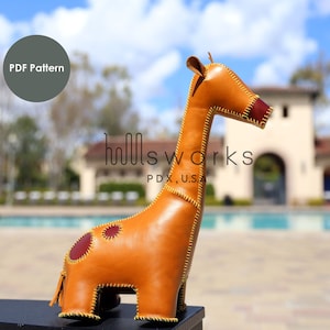May include: A tan leather giraffe toy with brown and red spots and yellow stitching. The giraffe is standing on a black surface with a blurred background of a swimming pool and a building. The text "hillsworks PDX, U.S.A." is printed on the giraffe. The text "PDF Pattern" is in a circle above the giraffe.