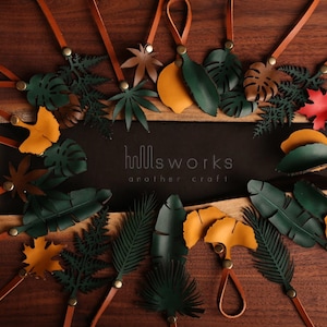 Pode incluir: Conjunto de pingentes de folhas de couro em v&aacute;rios tons de verde, marrom e amarelo. Os pingentes s&atilde;o presos a cord&otilde;es de couro marrom e est&atilde;o dispostos sobre uma superf&iacute;cie de madeira. O texto "hollsworks another craft" &eacute; vis&iacute;vel no centro da imagem.