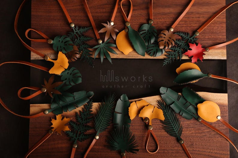 Pode incluir: Conjunto de pingentes de folhas de couro em v&aacute;rios tons de verde, marrom e amarelo. Os pingentes s&atilde;o presos a cord&otilde;es de couro marrom e est&atilde;o dispostos sobre uma superf&iacute;cie de madeira. O texto "hollsworks another craft" &eacute; vis&iacute;vel no centro da imagem.